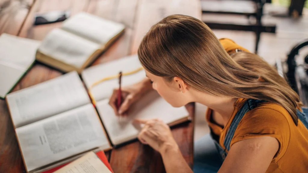 Student studying for SAT prep, writing notes in a notebook beside open textbooks.